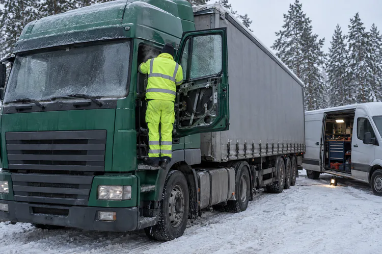 Zimą na leśnym parkingu mechanik naprawia podnośnik szyby w otwartych drzwiach zielonej ciężarówki; obok furgon serwisowy.