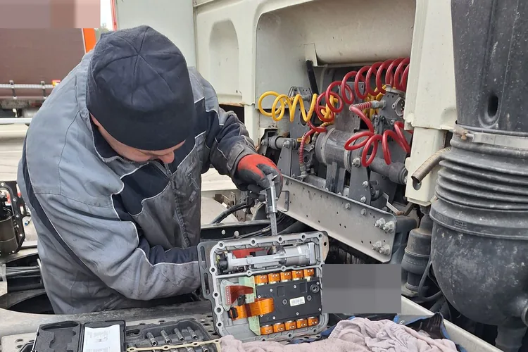 Mechanik w rękawicach pracuje przy module za kabiną ciągnika; na podeście rozłożone skrzynki z kluczami.