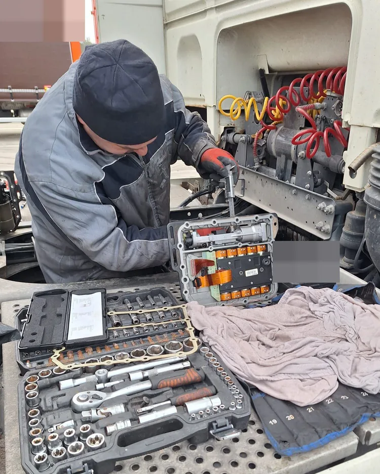Mechanik w rękawicach pracuje przy module za kabiną ciągnika; na podeście rozłożone skrzynki z kluczami.